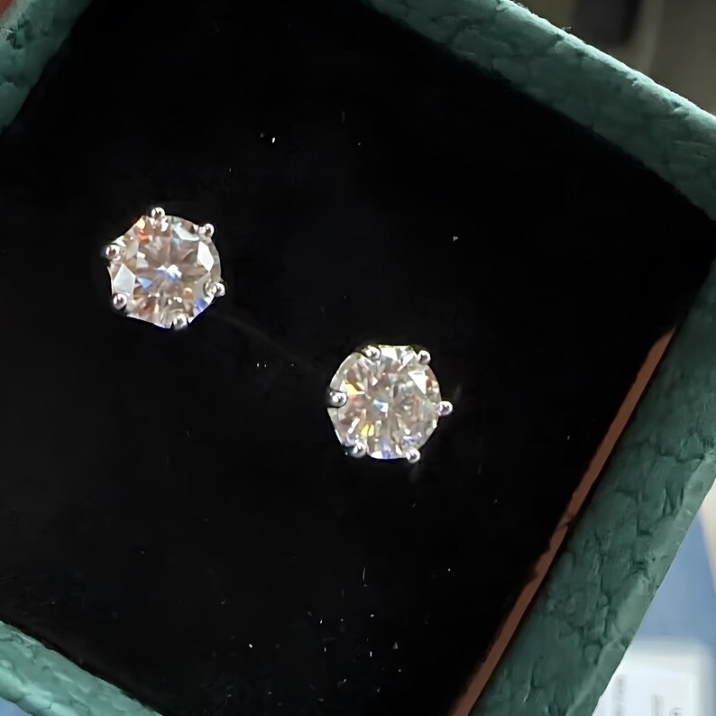 May include: A pair of sparkling diamond stud earrings, each with a round, brilliant-cut stone set in a silver or white gold setting. The earrings are displayed in a black jewelry box with a teal green exterior.