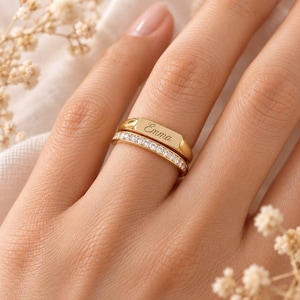Pode incluir: Um conjunto de dois anéis dourados sobrepostos. O anel superior apresenta uma placa retangular plana gravada com o nome Emma em letra cursiva. O anel inferior é incrustado com uma fileira de pequenas pedras claras e brilhantes.
