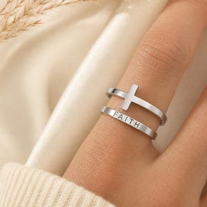 May include: A silver-toned double band ring featuring a cross symbol on the upper band and the word FAITH engraved in capital letters on the lower band. The ring has a polished metallic finish and a clean, open design that wraps around the finger.
