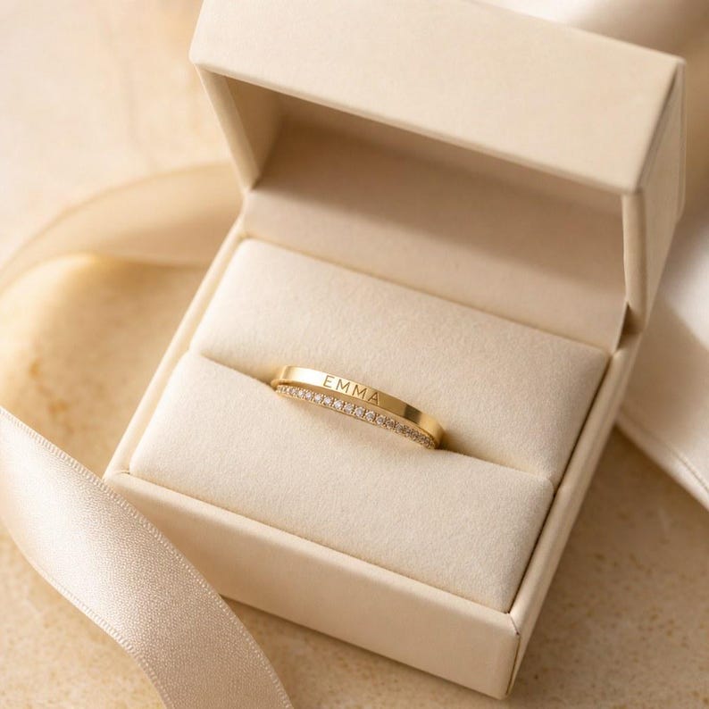May include: A gold band ring featuring the name EMMA engraved on the top surface and a row of small clear stones set along the lower edge, presented inside a cream-coloured jewellery box with a matching ribbon draped across the base.