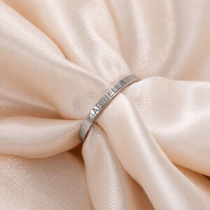 May include: A silver-toned metal ring with a smooth, flat band. The name GABRIELLA is engraved in capital letters along the outer surface of the band. The ring features a polished, reflective finish that catches the light across its curved surface.