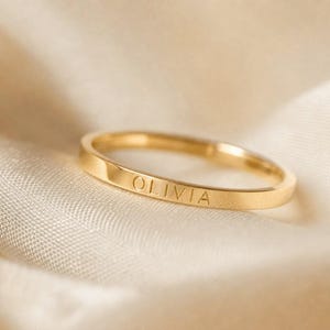 May include: A thin, polished gold band ring featuring the name OLIVIA engraved in clear, uppercase sans-serif lettering on the outer surface. The ring has a smooth, flat profile and a reflective metallic finish, showcasing a simple design.