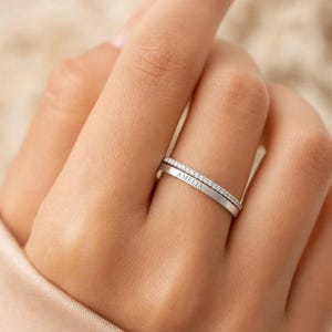 May include: A silver-toned double band ring featuring a thin upper band encrusted with small clear stones and a wider lower band engraved with the name AMELIA in capital letters. The two bands are connected to form a single piece of jewellery.