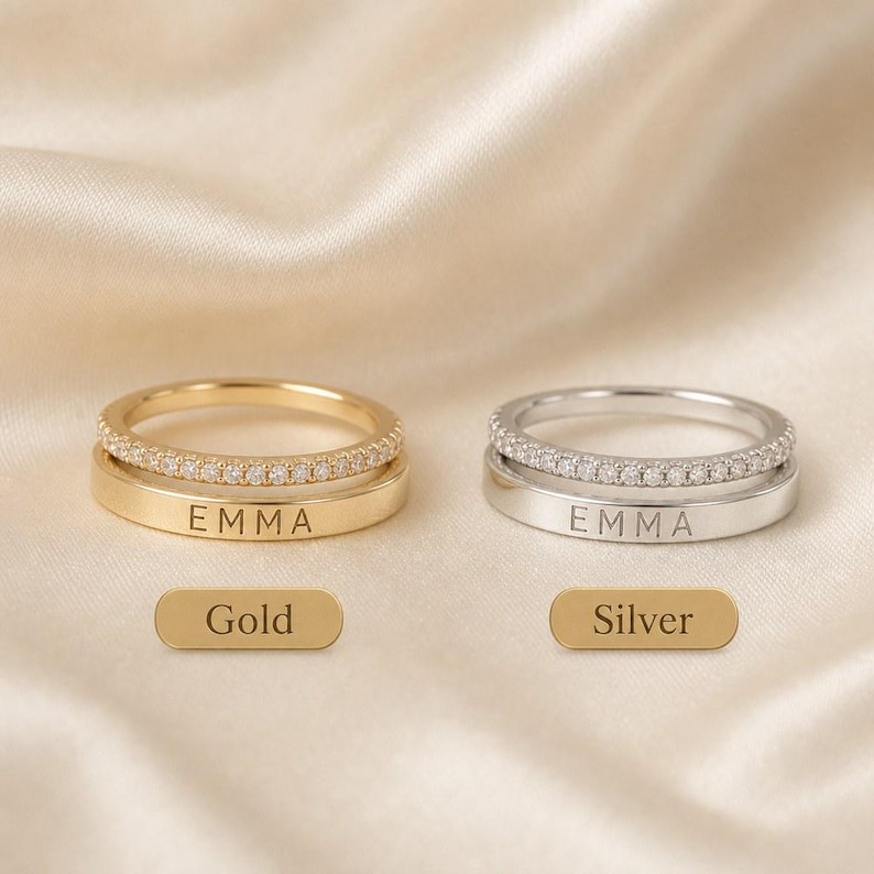 May include: Two rings, one gold and one silver, each featuring a smooth band engraved with the name EMMA and a second stacked band encrusted with small clear stones. Below the rings are two rectangular labels reading Gold and Silver.
