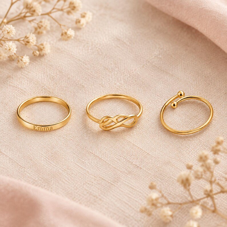 Pode incluir: Um conjunto de tr&ecirc;s an&eacute;is em tom dourado. O primeiro anel &eacute; uma banda simples gravada com o nome Emma. O segundo anel apresenta um design de s&iacute;mbolo de infinito. O terceiro anel &eacute; uma banda aberta com duas pequenas esferas nas pontas.