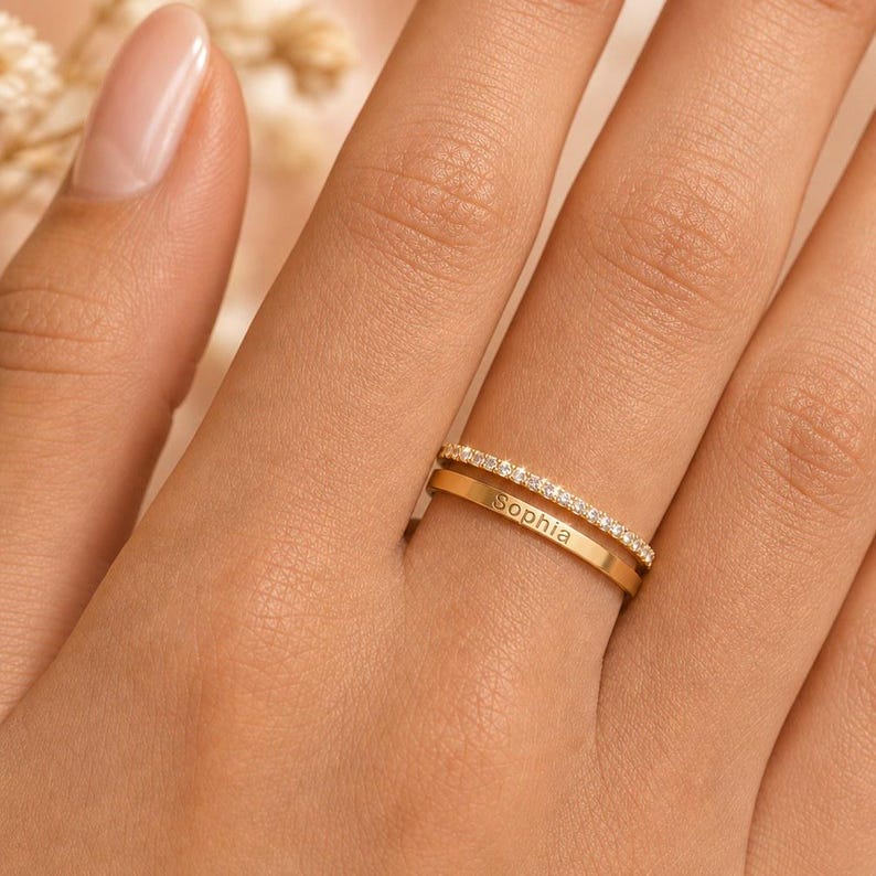 May include: A gold-toned double band ring featuring one band set with a row of small clear stones and a second plain band engraved with the name Sophia. The two bands are connected to form a single piece of jewellery with a polished metallic finish.