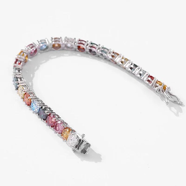 May include: A silver bracelet with a row of colorful gemstones. The gemstones are arranged in a rainbow pattern, with a variety of colors including red, orange, yellow, green, blue, and purple. The bracelet has a clasp closure.