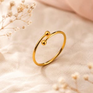 Pode incluir: Um anel aberto em tom dourado com uma banda fina e polida que termina em dois pequenos detalhes esf&eacute;ricos nas extremidades. O metal tem uma superf&iacute;cie lisa e reflexiva, e o design caracteriza-se pela sua estrutura circular, simples e ajust&aacute;vel.