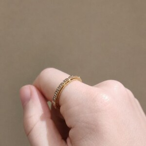 May include: A gold-toned ring featuring a thin band encrusted with a single row of small, clear, sparkling stones. The ring has a polished metallic finish and a slender profile, showcasing a continuous line of light-reflecting gems along the outer surface.