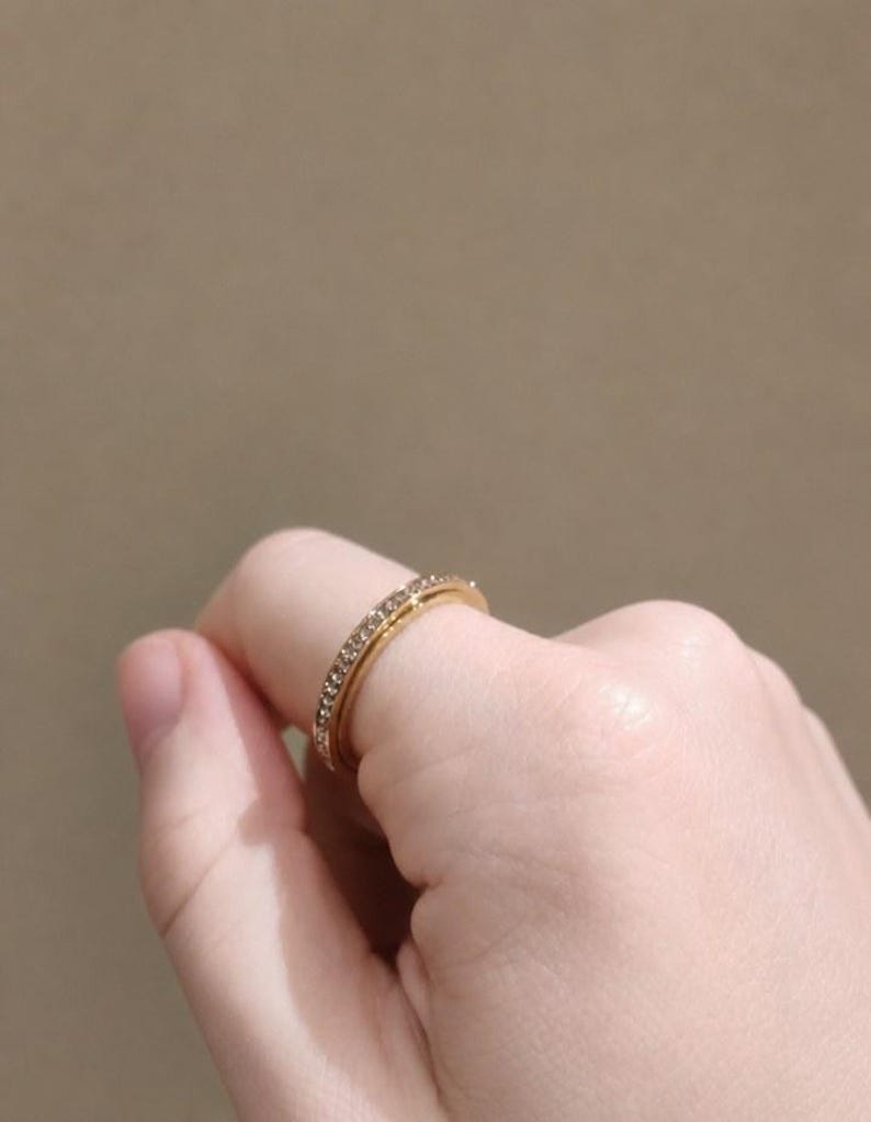 May include: A gold-toned ring featuring a thin band encrusted with a single row of small, clear, sparkling stones. The ring has a polished metallic finish and a slender profile, showcasing a continuous line of light-reflecting gems along the outer surface.