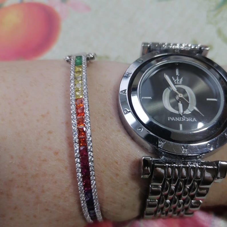 May include: A silver-toned Pandora watch with a dark gray face and a matching bracelet. The bracelet features a rainbow of colorful gemstones. The watch and bracelet are worn on a wrist.