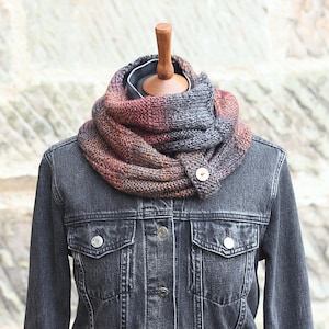 May include: A hand-knitted cowl scarf in shades of gray, red, and brown, fastened with a wooden button. The scarf is displayed on a mannequin, worn over a dark denim jacket.