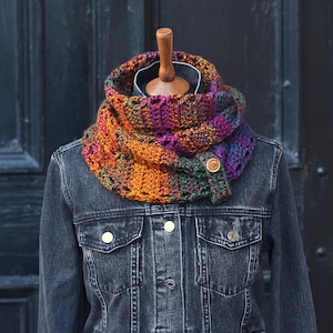May include: A colorful, hand-knitted cowl scarf in shades of orange, purple, and green. The scarf is buttoned with a wooden button and is displayed on a mannequin wearing a denim jacket. The cowl is a cozy accessory for cold weather.