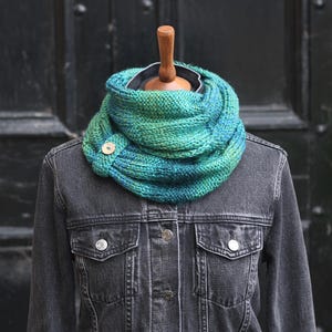 May include: A teal and green knitted cowl with a wooden button fastening, displayed on a mannequin wearing a dark denim jacket. The cowl's textured knit design adds a handmade feel. The cowl is a stylish accessory.