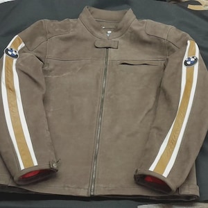 May include: A brown suede jacket with a full-length zip, a small collar, and a chest pocket. The sleeves have white and gold stripes and a BMW logo patch. The jacket has red lining at the cuffs.