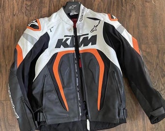 Puede incluir: Chaqueta de moto negra, blanca y naranja con el logo de KTM. La chaqueta tiene cremallera frontal, hombros acolchados y cuello alto. La chaqueta tiene un exterior de cuero y está colgada de una percha.