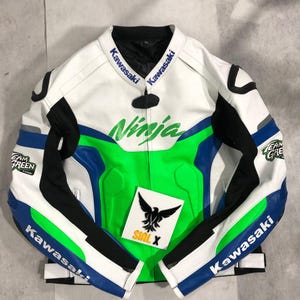 Könnte beinhalten: Weiße, schwarze, grüne und blaue Kawasaki Ninja Motorradjacke. Die Jacke hat das Wort "Ninja" in Grün auf der Brust, mit "Kawasaki" auf Kragen und Ärmeln. Ein schwarz-weißes Logo befindet sich auf der Brust.