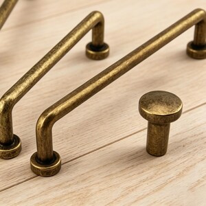 May include: Close-up of antique brass cabinet hardware, including three bar pulls and one knob. The hardware has a warm, aged finish and is set against a light wood surface. The pulls have a curved design.