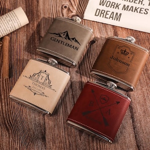 May include: Four personalised leather-wrapped hip flasks with different designs. The flasks are brown, beige, red, and grey. The designs include a mountain range, a crest with a crown, a crest with an initial, and a crossed arrow design.