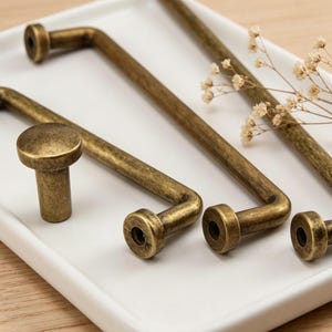 Op de afbeelding: Close-up van antieke messing kastbeslagen, waaronder twee L-vormige handgrepen, een rechte stanggreep en een ronde knop. De hardware is gerangschikt op een wit dienblad met gedroogde bloemen.