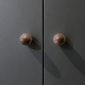Pu&ograve; includere: Due pomelli rotondi in legno con un motivo a venature a strisce, montati su un'anta di un armadio grigio scuro. I pomelli sono rotondi e hanno una finitura in legno naturale. Le ante dell'armadio sono grigie opache.