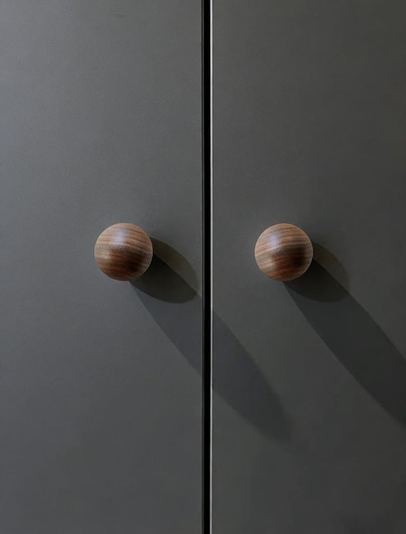 Pu&ograve; includere: Due pomelli rotondi in legno con un motivo a venature a strisce, montati su un'anta di un armadio grigio scuro. I pomelli sono rotondi e hanno una finitura in legno naturale. Le ante dell'armadio sono grigie opache.