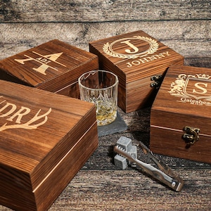 Op de afbeelding: Vier houten kisten met gegraveerde ontwerpen en gepersonaliseerde tekst. Eén kist heeft de naam "JOHNNY" in een lauwerkrans. Een glas amberkleurige vloeistof, ijsblokjes en een tang zijn ook aanwezig.