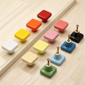 Könnte beinhalten: Eine Sammlung quadratischer Schrankknöpfe in verschiedenen Farben, darunter Rot, Orange, Gelb, Rosa, Blau, Grün und Weiß. Jeder Knopf hat eine glänzende Oberfläche und eine Messingschraube zur Befestigung. Die Knöpfe sind auf einer hellen Holzoberfläche angeordnet.