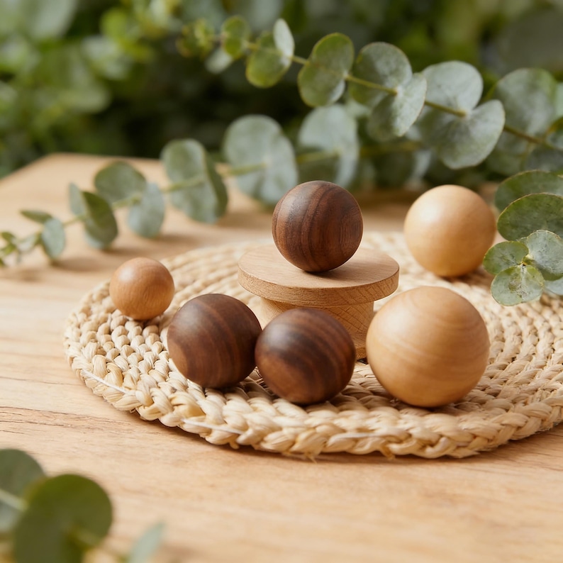 Pu&ograve; includere: Una collezione di sfere di legno in varie dimensioni e tonalit&agrave; di marrone e beige. Alcune sfere sono in noce scuro, altre in acero chiaro. Le sfere sono disposte su un tappetino intrecciato e un piccolo piedistallo di legno, con foglie di eucalipto sullo sfondo.