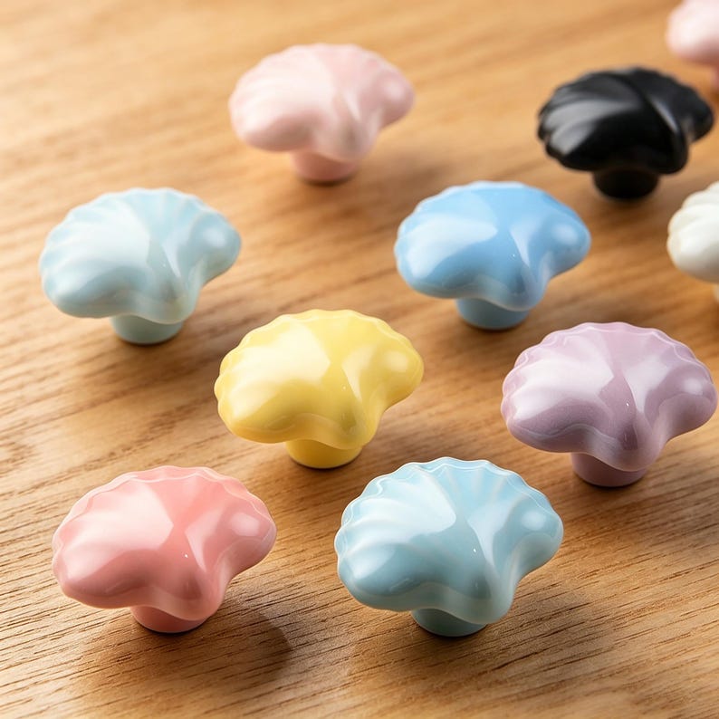 K&ouml;nnte beinhalten: Eine Sammlung von muschelf&ouml;rmigen Keramik-Schrankkn&ouml;pfen in verschiedenen Pastellfarben, darunter Rosa, Blau, Gelb und Lila. Die Kn&ouml;pfe haben eine gl&auml;nzende Oberfl&auml;che und liegen auf einer Holzoberfl&auml;che.