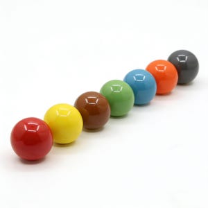 May include: A row of seven colorful, glossy, round cabinet knobs. The knobs are in a gradient of colors: red, yellow, brown, green, blue, orange, and gray. Each knob has a small, square indentation on the top. The knobs are set against a white background.