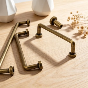 May include: Antique brass cabinet pulls and knobs. The hardware is a warm, aged brass color. The pulls are rectangular with rounded corners, and the knobs are cylindrical. The hardware is displayed on a light wood surface.