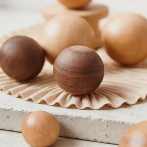 Pu&ograve; includere: Sfera di legno in varie tonalit&agrave; di marrone e beige. Le sfere sono disposte su una superficie beige a pieghe e su una base di cemento. La venatura del legno &egrave; visibile su alcune sfere, evidenziando le variazioni naturali di consistenza e colore.