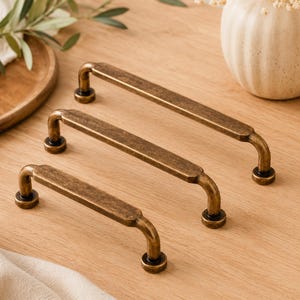May include: Three antique brass cabinet pulls with a curved handle design. The pulls are in varying sizes and are set on a light wood surface. The hardware has a vintage aesthetic.