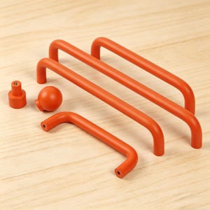 May include: A collection of orange cabinet hardware, including three long, curved handles, a smaller handle, a round knob, and a cylindrical knob. The hardware is made of a solid material and is displayed on a light wood surface.