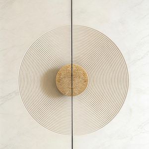 May include: A cabinet door with a cream-coloured, concentric circle design. The door has a gold-coloured, textured knob at the centre. The background is a light marble pattern. The door is split by a black line.
