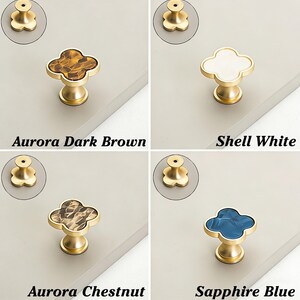 K&ouml;nnte beinhalten: Vier dekorative Schrankkn&ouml;pfe mit goldfarbenem Sockel und Kleeblatt-Design. Die Farben sind Aurora Dark Brown, Shell White, Aurora Chestnut und Sapphire Blue. Der Text auf dem Bild lautet "Aurora Dark Brown", "Shell White", "Aurora Chestnut" und "Sapphire Blue".