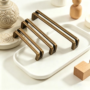 May include: Three bronze-coloured cabinet pulls of varying sizes are arranged on a white tray. The pulls have a rectangular design with a double bar. The pulls are made of metal and have a vintage aesthetic. The tray is surrounded by other decorative items.