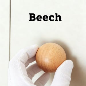 Pu&ograve; includere: Una sfera di legno marrone chiaro tenuta da una mano guantata. La parola "Beech" &egrave; stampata in grassetto nero sopra la sfera. La sfera &egrave; fatta di legno e ha una superficie liscia e lucida.