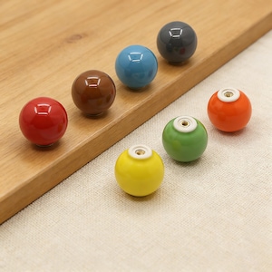 Può includere: Una collezione di pomelli per mobili in ceramica colorata in varie tonalità, tra cui rosso, marrone, blu, grigio, arancione, verde e giallo. Ogni pomello è sferico con una piccola rientranza quadrata e una base bianca.