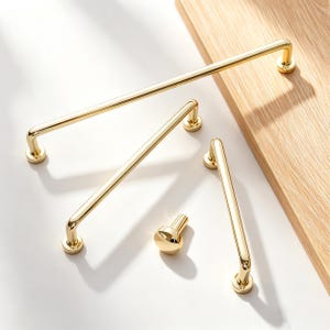 May include: A collection of gold-colored cabinet hardware, including several bar pulls and a knob. The hardware is displayed on a white surface with a wooden cutting board in the background. The pulls have a sleek, modern design.
