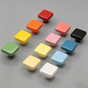 May include: A collection of square cabinet knobs in various glossy colors, including red, orange, yellow, green, pink, black, blue, and white. The knobs are arranged on a gray surface, showcasing their vibrant hues and smooth finish.