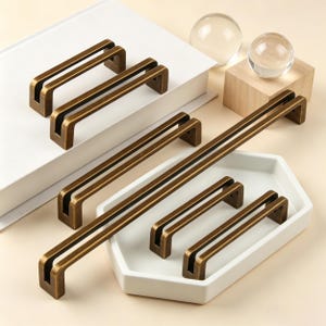 May include: Bronze-coloured cabinet pulls in various sizes are displayed on a white surface and a white tray. The pulls have a rectangular design with a double bar. Two clear glass spheres and a wooden cube are also in the image.