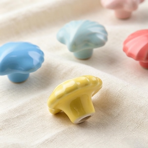 K&ouml;nnte beinhalten: Eine Sammlung farbenfroher, pilzf&ouml;rmiger Keramik-Schrankkn&ouml;pfe in Blau-, Gelb- und Rosat&ouml;nen. Die Kn&ouml;pfe sind auf einem cremefarbenen Stoff angeordnet, der ihre gl&auml;nzende Oberfl&auml;che und ihr einzigartiges Design zur Geltung bringt. Diese dekorativen Kn&ouml;pfe verleihen M&ouml;beln einen skurrilen Touch.
