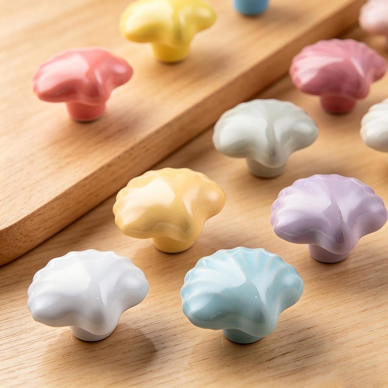 K&ouml;nnte beinhalten: Eine Sammlung farbenfroher, pilzf&ouml;rmiger Schrankkn&ouml;pfe aus Keramik. Die Kn&ouml;pfe sind in verschiedenen Pastellfarben wie Rosa, Gelb, Blau und Wei&szlig; gehalten. Sie sind auf einer Holzoberfl&auml;che angeordnet und zeigen ihr dekoratives Design.