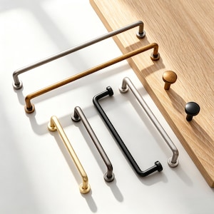 May include: A collection of cabinet hardware in various metallic finishes, including silver, gold, and black. The assortment features bar pulls and knob styles, displayed on a wooden surface. The hardware is designed for furniture and cabinetry.