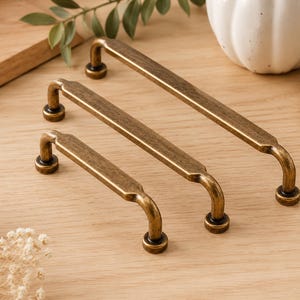 May include: Three antique brass cabinet pulls of different sizes are displayed on a light wood surface. The pulls feature a rectangular bar design with curved ends and circular mounting bases. Decorative furniture hardware.