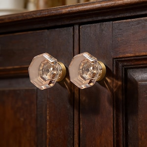 May include: Two clear, faceted glass cabinet knobs with a soft pink hue, mounted on brass bases. The knobs are octagonal and attached to a dark brown wooden cabinet. The hardware adds a touch of elegance to the furniture.