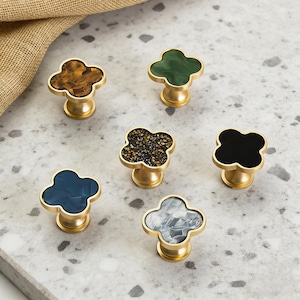 K&ouml;nnte beinhalten: Sechs dekorative Schrankkn&ouml;pfe mit Kleeblatt-Design. Die Kn&ouml;pfe haben einen goldfarbenen Sockel und weisen verschiedene Farben und Muster auf, darunter Braun, Gr&uuml;n, Schwarz, Blau und Grau. Die Kn&ouml;pfe sind auf einer hellgrauen Oberfl&auml;che angeordnet.