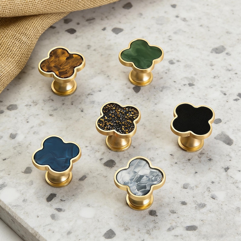 K&ouml;nnte beinhalten: Sechs dekorative Schrankkn&ouml;pfe mit Kleeblatt-Design. Die Kn&ouml;pfe haben einen goldfarbenen Sockel und weisen verschiedene Farben und Muster auf, darunter Braun, Gr&uuml;n, Schwarz, Blau und Grau. Die Kn&ouml;pfe sind auf einer hellgrauen Oberfl&auml;che angeordnet.